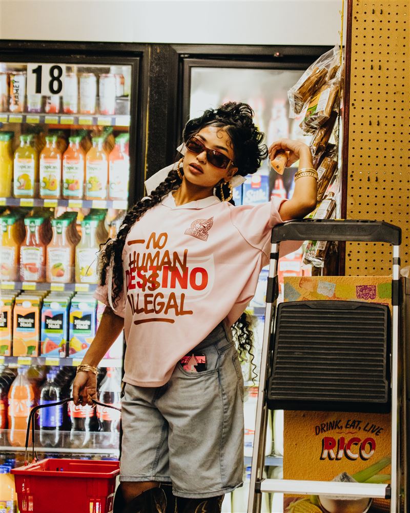 Woman in the pink Destino jersey and sunglasses leaning on a ladder inside a mercado, warm store light