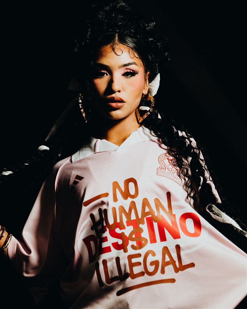 Woman in a pink Destino jersey reading No Human Is Illegal, lit by a single small softbox against black